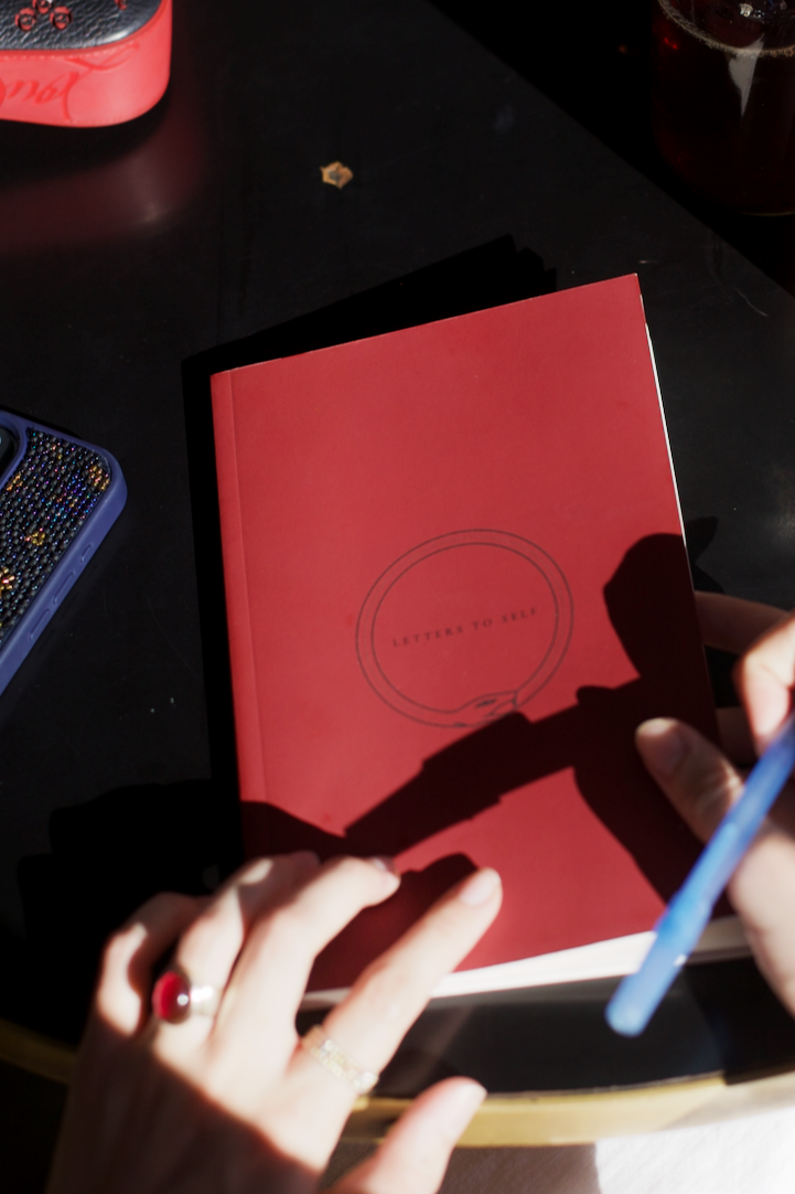 Letters To Self red guided journal on a dark surface, with hands holding a pen.
